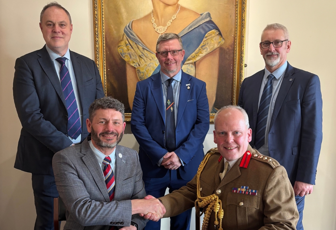 Two people shake hands, one in a suit and one in military uniform, with three others standing behind them beneath a framed portrait.